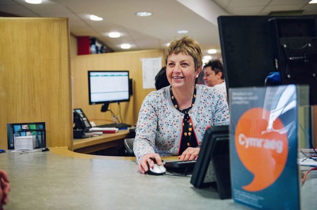 Dynes yn eistedd wrth ddesg gyda poster Iaith Gwaith